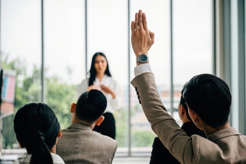 In a seminar classroom at a conference, a large group raises hands in participation. The engaged